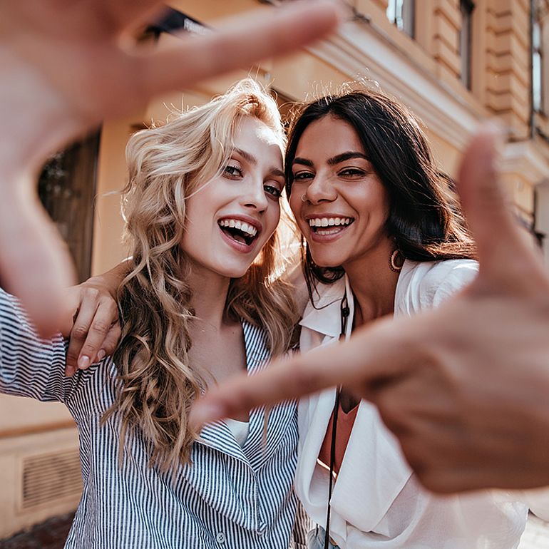 Due donne sorridono e con le dita formano una cornice attorno ai loro volti.