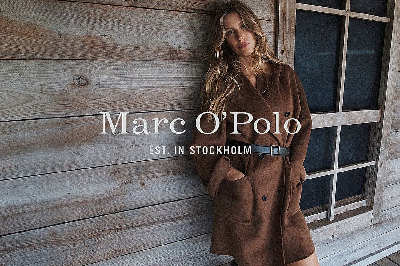 Una donna è appoggiata al muro di una casa, con una finestra alle sue spalle, e indossa un cappotto.