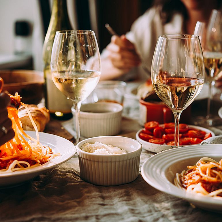 Una tavola imbandita; sopra di essa, due persone brindano con un bicchiere di vino ciascuno.