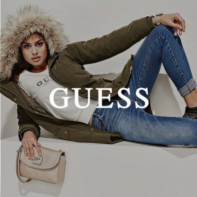 Una donna è sdraiata di lato sul divano, con un ginocchio piegato e una mano appoggiata su di esso. Nell'altra mano tiene una borsa. Sopra di essa è posizionato il logo Guess.