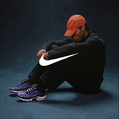 Un uomo è seduto per terra con entrambe le ginocchia piegate verso il corpo. Il suo sguardo è rivolto verso il pavimento. Sopra di lui è posizionato il logo Nike.