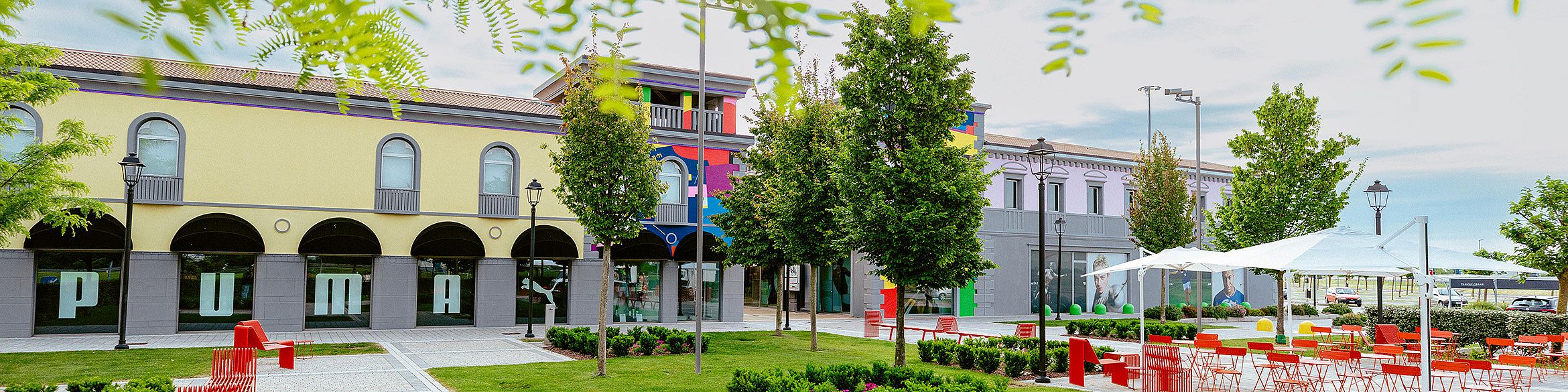 Viene mostrata l'entrata del Palmanova Designer Village. È visibile il negozio Puma. Davanti all'entrata ci sono tavoli e sedie.